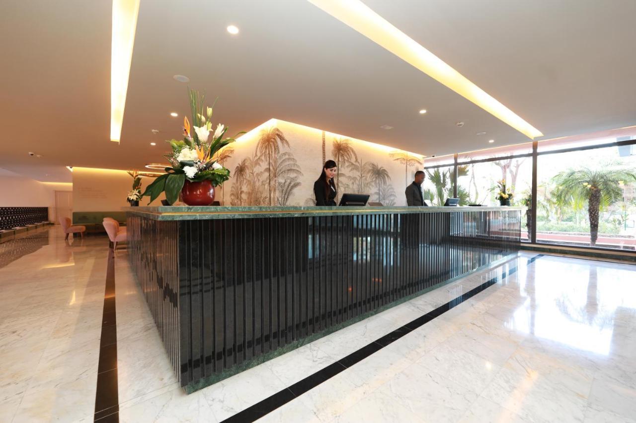 Hotel Kenzi Rose Garden Marrakesh Exterior foto The image shows a modern reception area, likely for a hotel or office building. There is a stylish front desk made of dark materials with a smooth surface. Behind the desk, two staff members are engaged in their work, possibly checking in guests or a